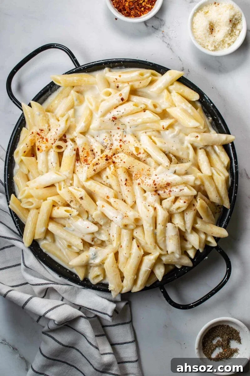 Top down view of a black pan of creamy pasta on a white backdrop with cheese and chili flakes in small white bowls.