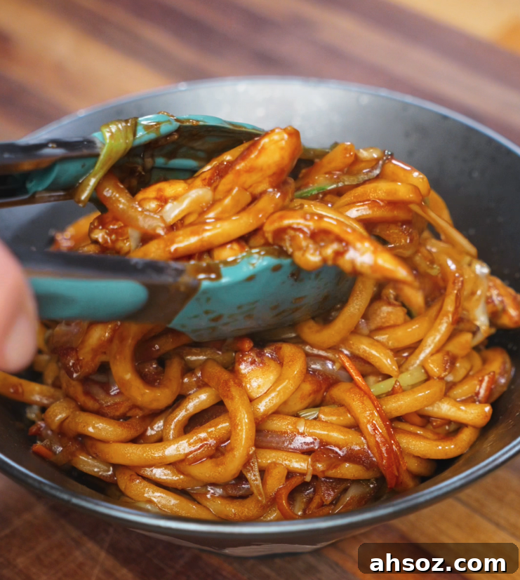 A pair of teal tongs places a serving of glossy chicken yaki udon into a black bowl. The chewy noodles, stir-fried with tender chicken, onions, and carrots, are coated in a rich soy-based sauce.