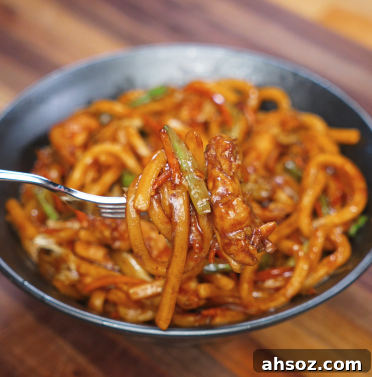 A fork lifts a saucy bite of chicken yaki udon with glistening noodles and stir-fried vegetables, showing the glossy texture of the stir-fry.