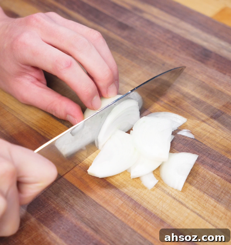A hand uses a chef’s knife to thinly slice half an onion on a wooden cutting board for the vegetable base of the stir-fry.