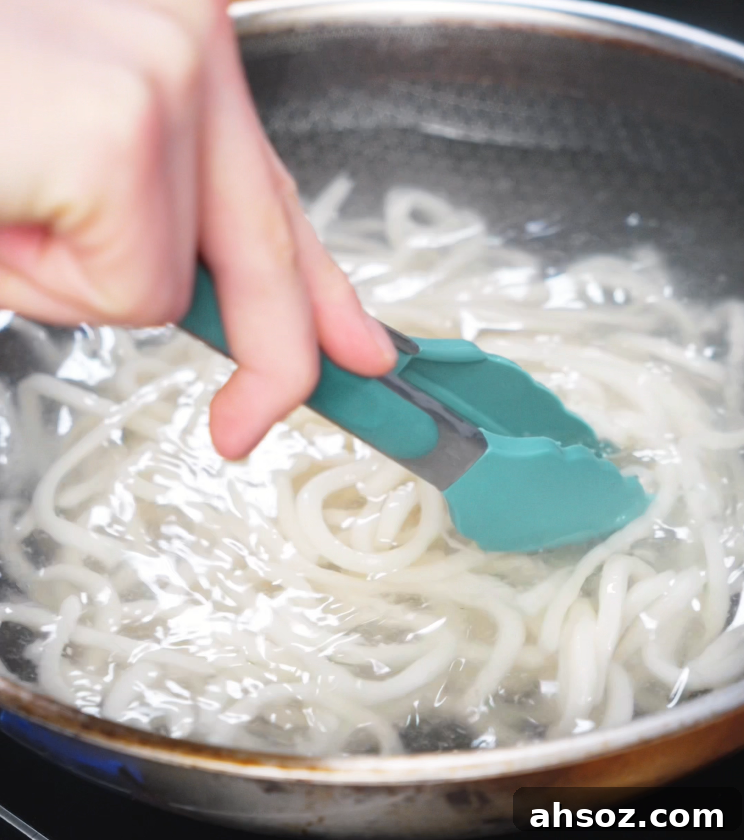 A pair of teal tongs stirs thick udon noodles in boiling water to prepare them for the chicken yaki udon stir-fry.