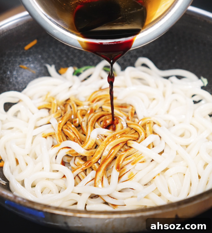 A soy-based sauce is poured over freshly boiled udon noodles in a pan filled with stir-fried veggies, ready to be tossed with chicken.