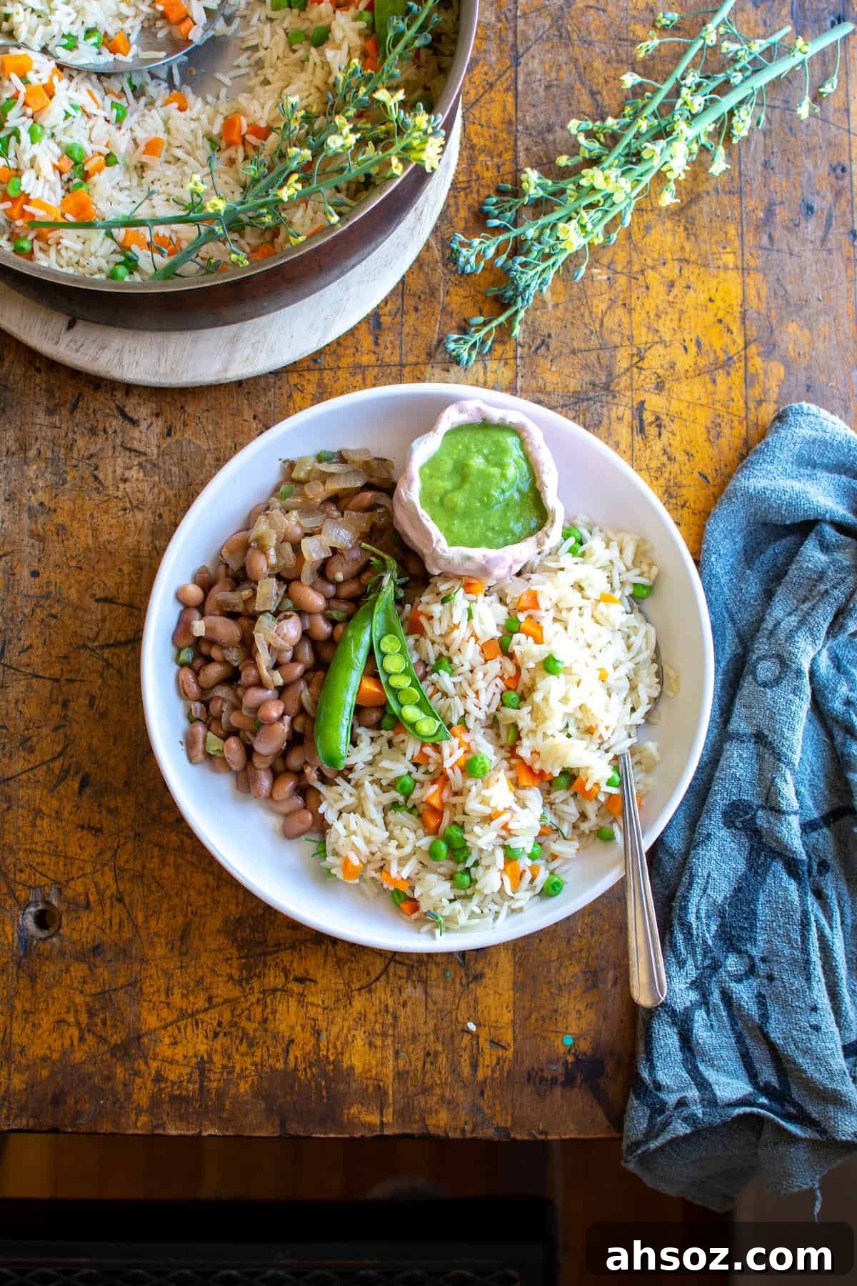A white plate filled with rice and beans sitting on a wood table. There is a pot of Arroz behind the plate and a napkin on the side. 