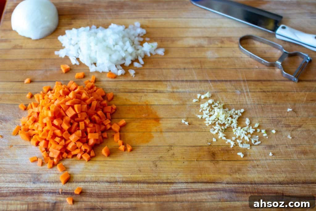 Diced carrots, white onion, and minced garlic on a wood cutting board with the other half of the onion behind them. 