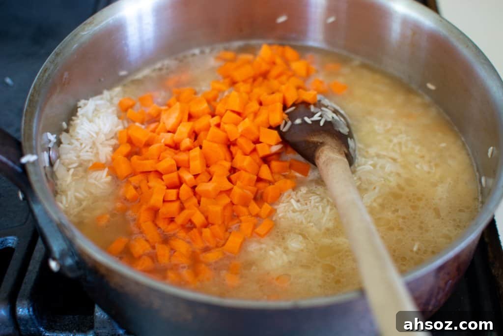 A large pot of rice with diced carrots and chicken broth sitting on the stove with a wood spoon sticking in one side. 