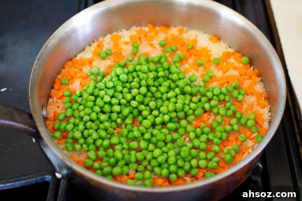 A pot with cooked white rice and carrots and a pile of frozen peas on top sitting on the stove. 