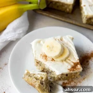 A banana bar with cream cheese icing on a plate, with bananas and more bars in the background.