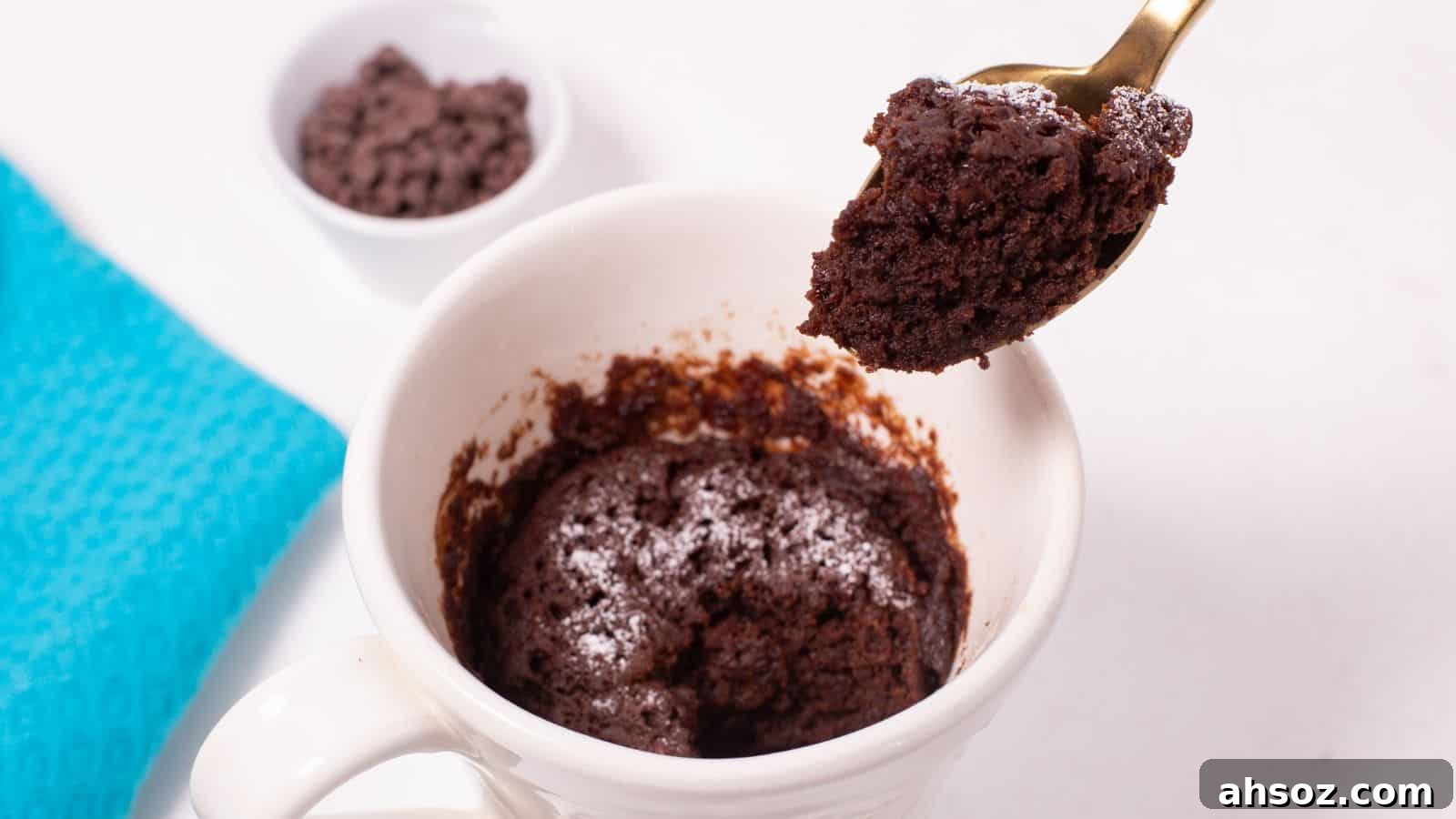 A spoon lifting a bite of freshly made chocolate mug cake from a white mug, with chocolate chips in a small bowl in the background.