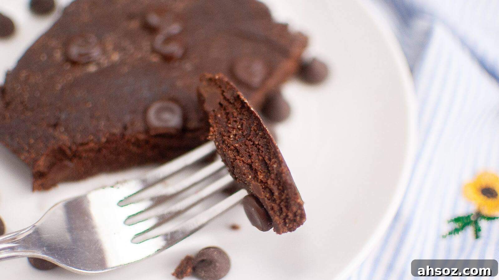 Fork holding a slice of a chocolate brownie with chocolate chips on a white plate.