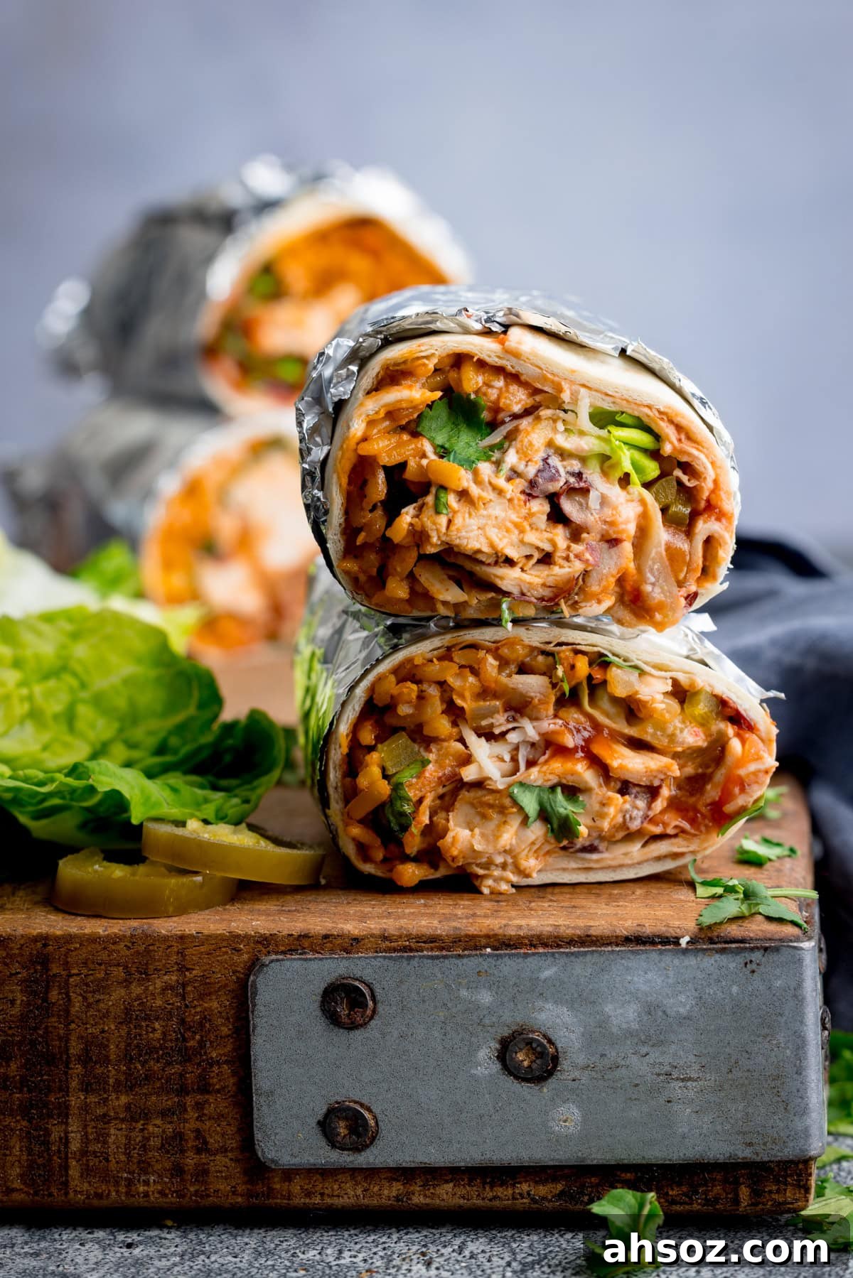Two foil-wrapped rotisserie chicken burritos are stacked on a wooden board, revealing fillings of chicken, rice, beans, and vegetables. Lettuce and sliced jalapeños are next to the burritos on the board.