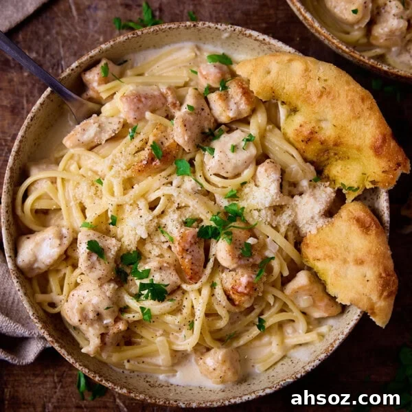 A stoneware bowl on a wooden board, the bowl is full of garlic parmesan chicken pasta with garlic bread and a fork.