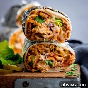 Two halves of a rotisserie chicken burrito packed with rice, rotisserie chicken, cheese, beans, and vegetables are wrapped in foil and garnished with coriander. They sit stacked on a wooden surface.