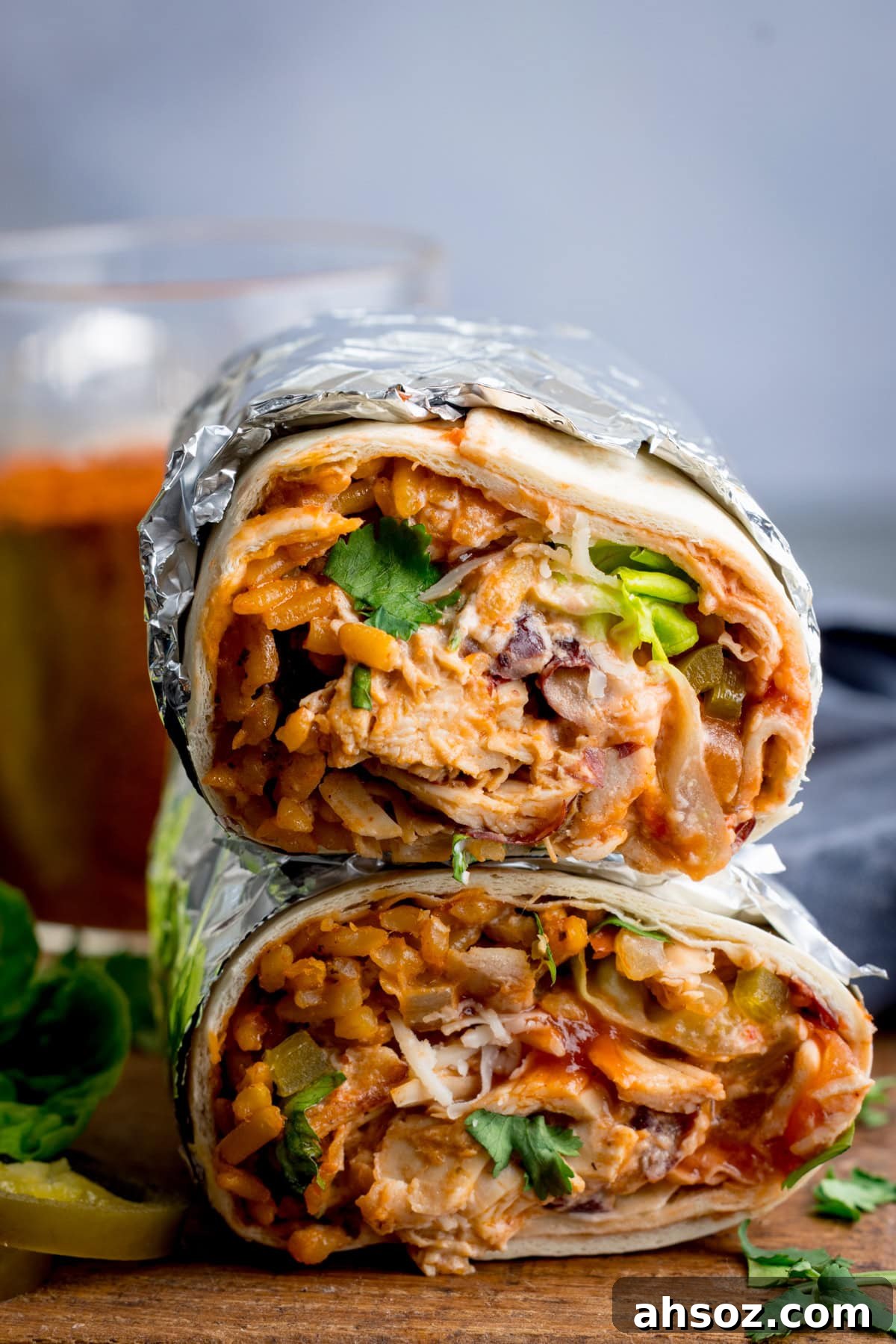 A rotisserie chicken burrito cut in half reveals layers of rice, cheese, beans, and coriander wrapped in a flour tortilla with aluminium foil.