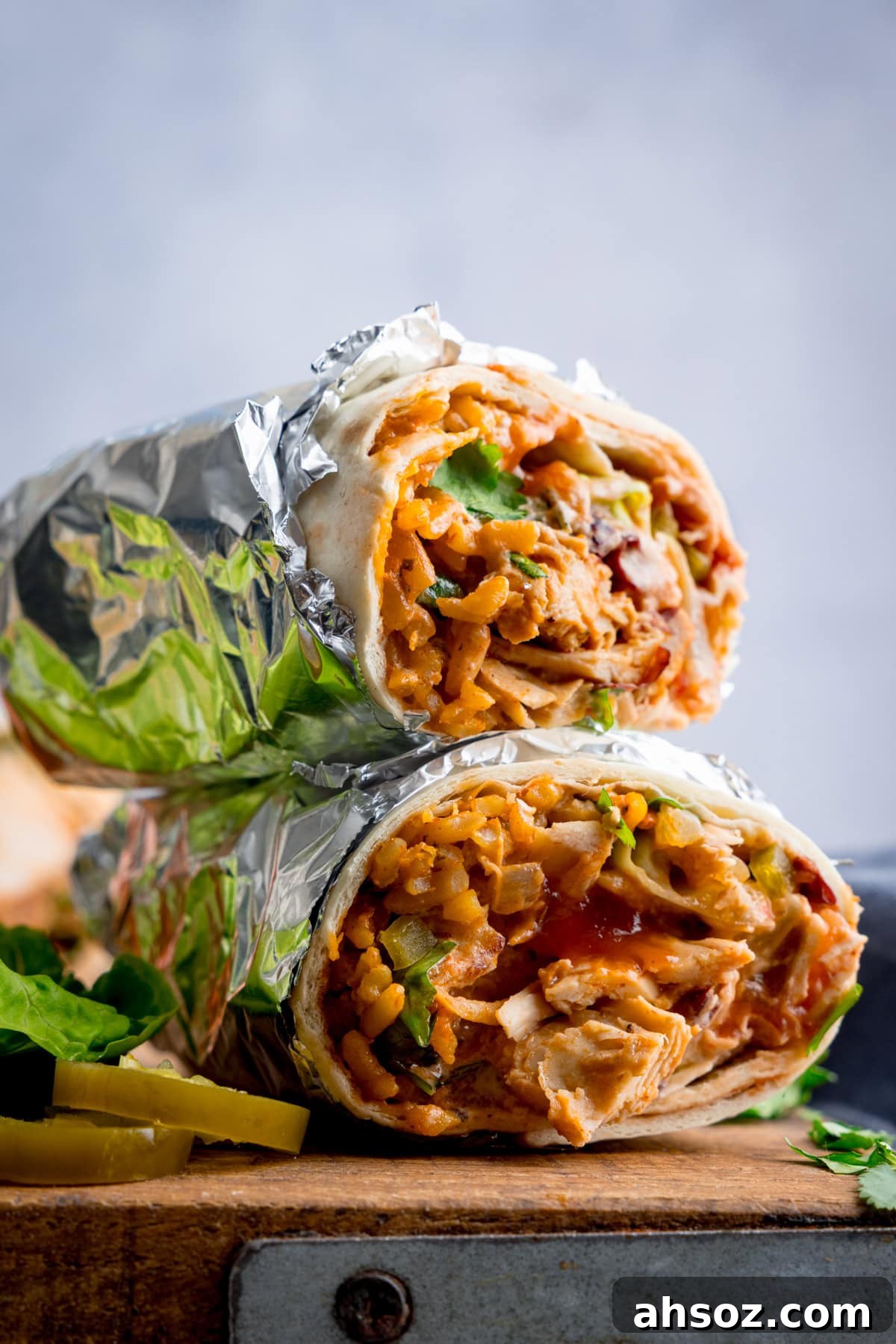 Two foil-wrapped Rotisserie Chicken Burrito halves stacked, revealing fillings of rice, chicken, vegetables, and sauce, on a wooden board. There are a couple of slices of jalapeno next to the burrito.