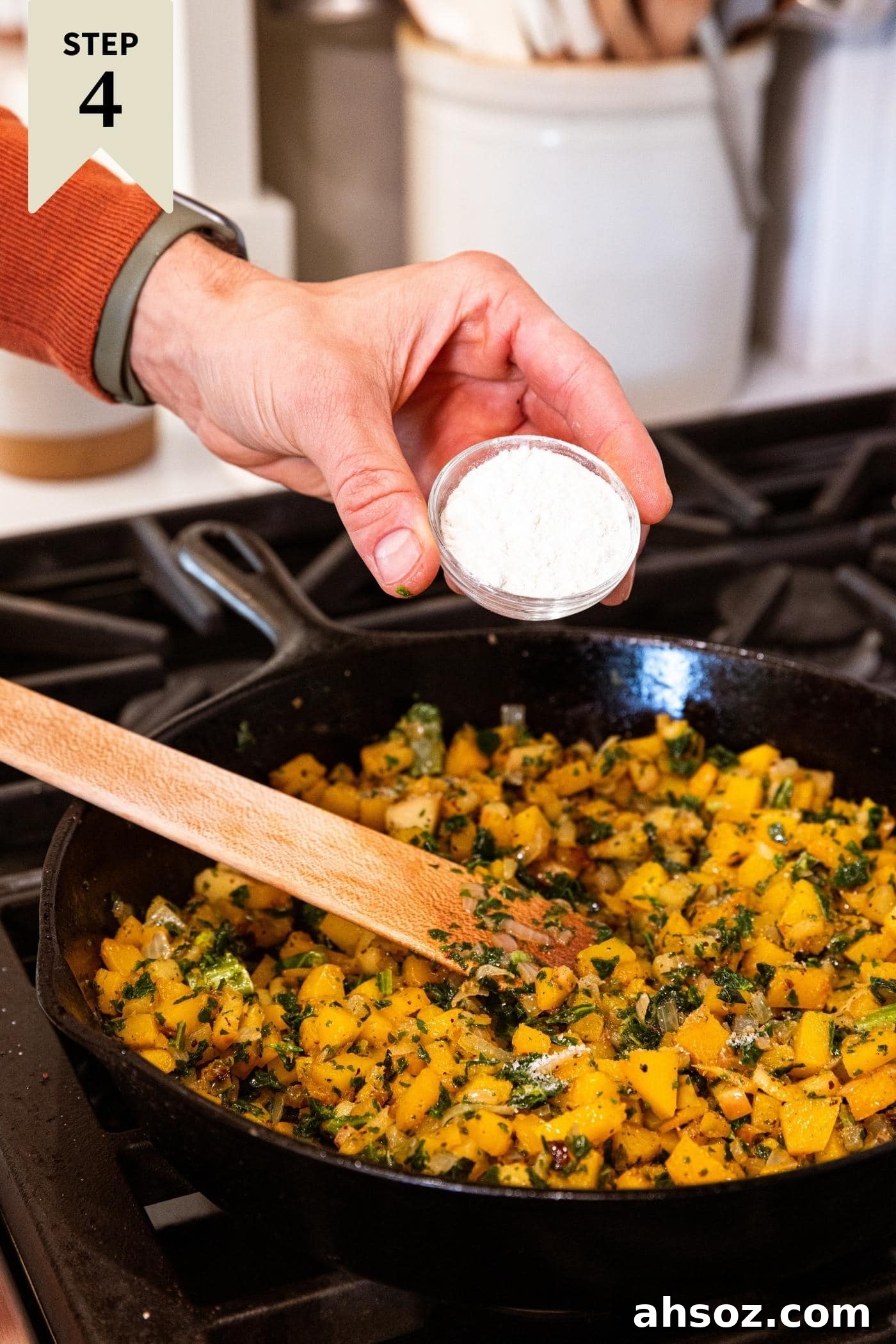 Hand holding small glass bowl filled with flour about to pour into a black cast iron skillet filled with butternut squash and kale mixture on the stove after cooking for some time.