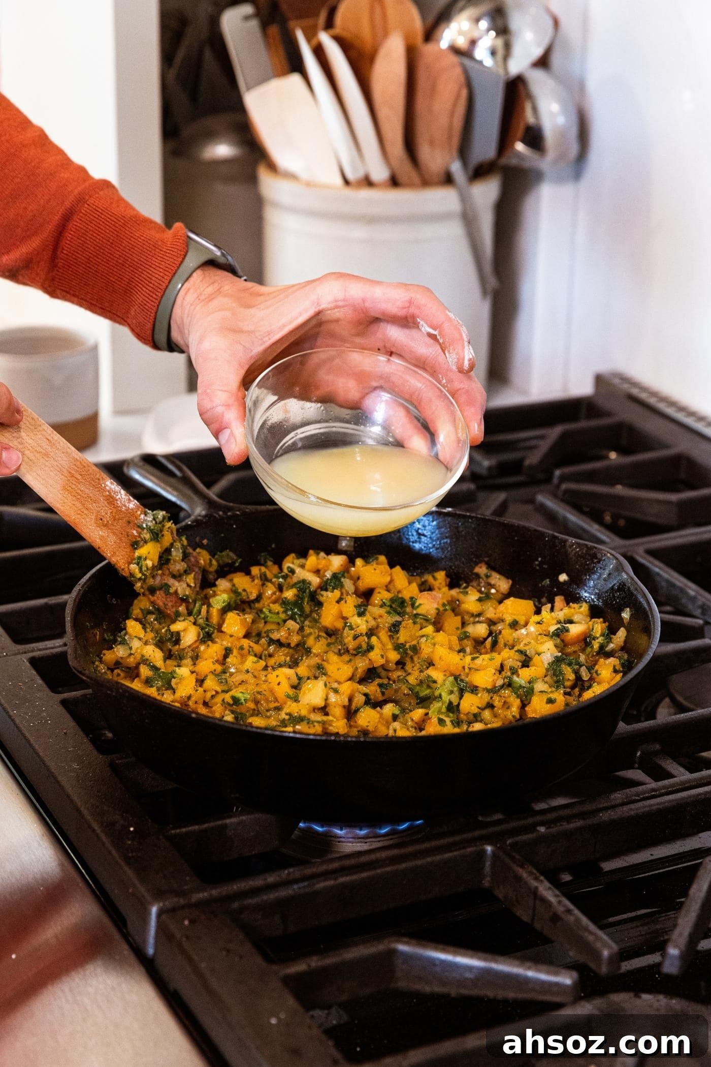 Hand holding glass bowl filled with light yellow colored chicken stock about to pour into cast iron skillet filled with diced butternut squash and kale.