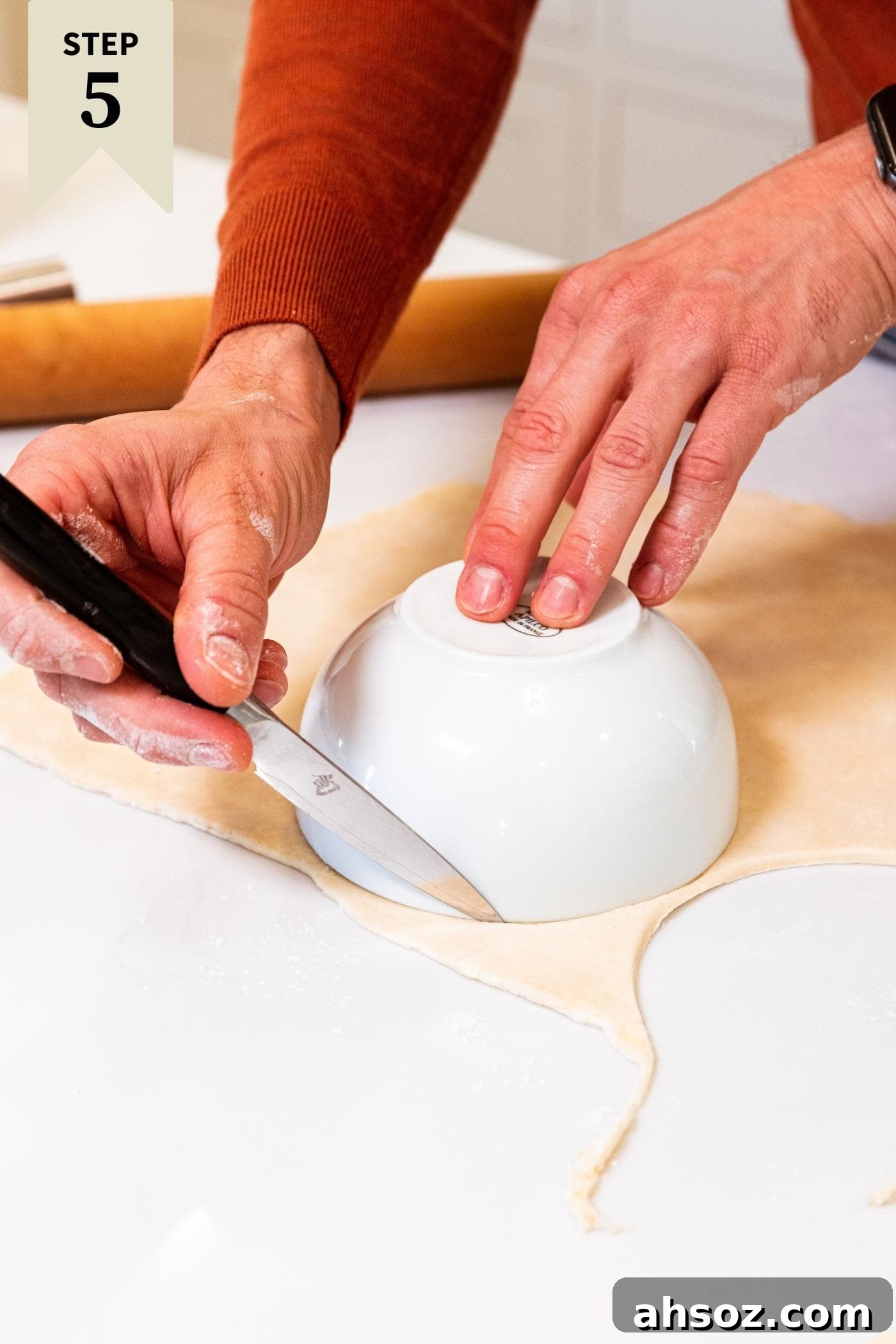 Hand using knife to cut out circle from dough using a white bowl laid upside down on top of dough all on white countertop.