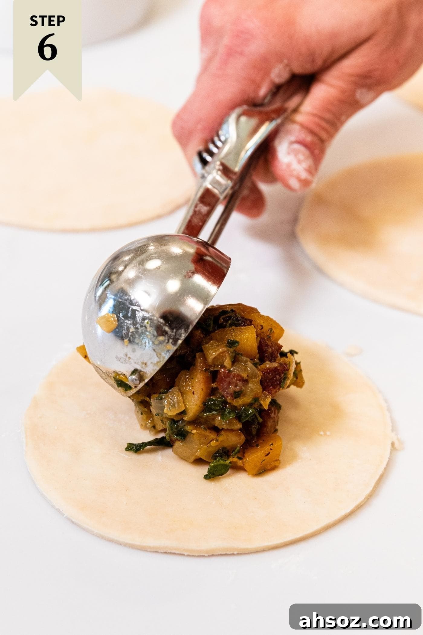 Hand using metal cookie scoop to fill up a round of dough with butternut squash and sausage mixture on white countertop.