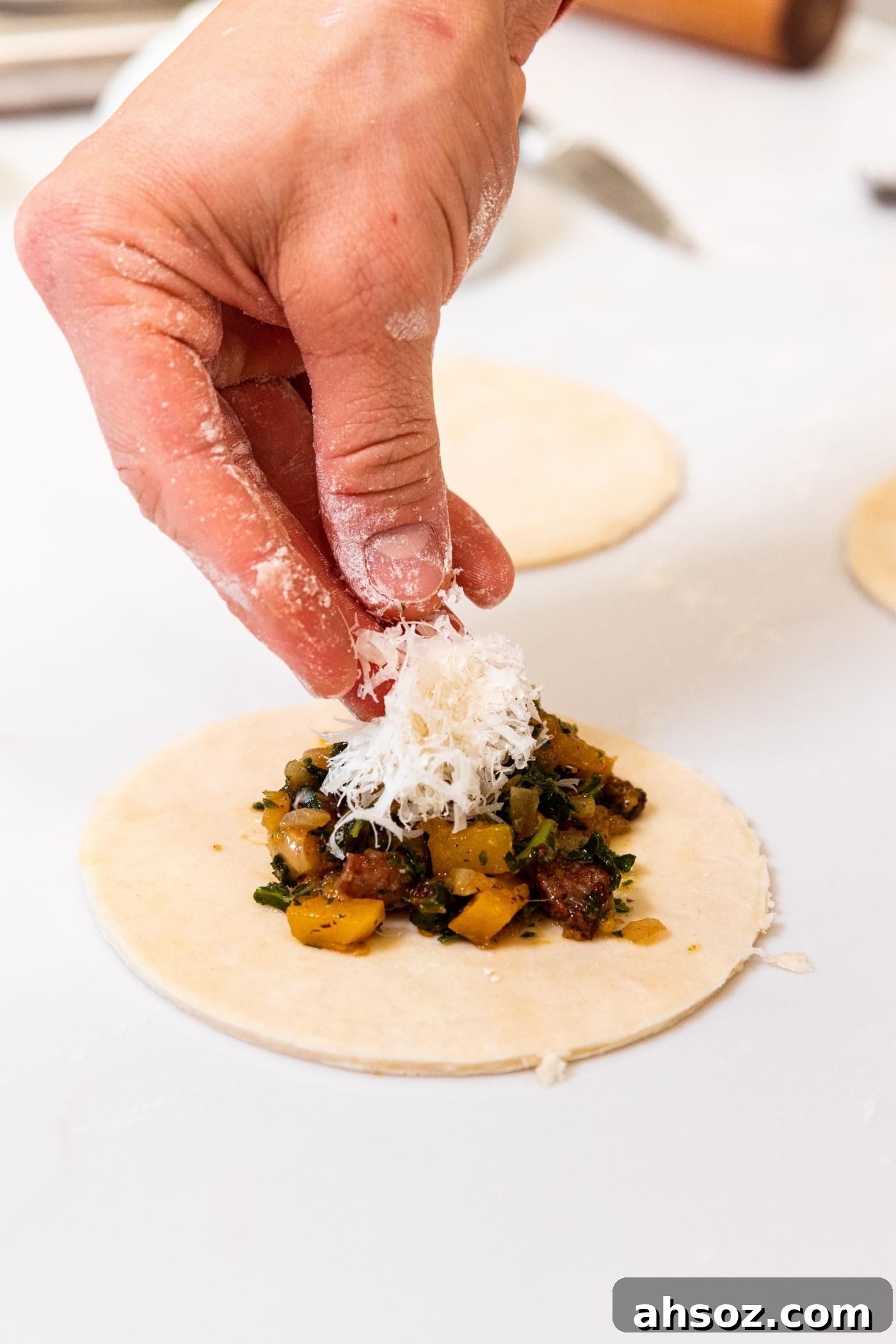 Hand dropping a sprinkling of Parmesan cheese on top of butternut squash mixture on top of round of pie dough on white countertop.