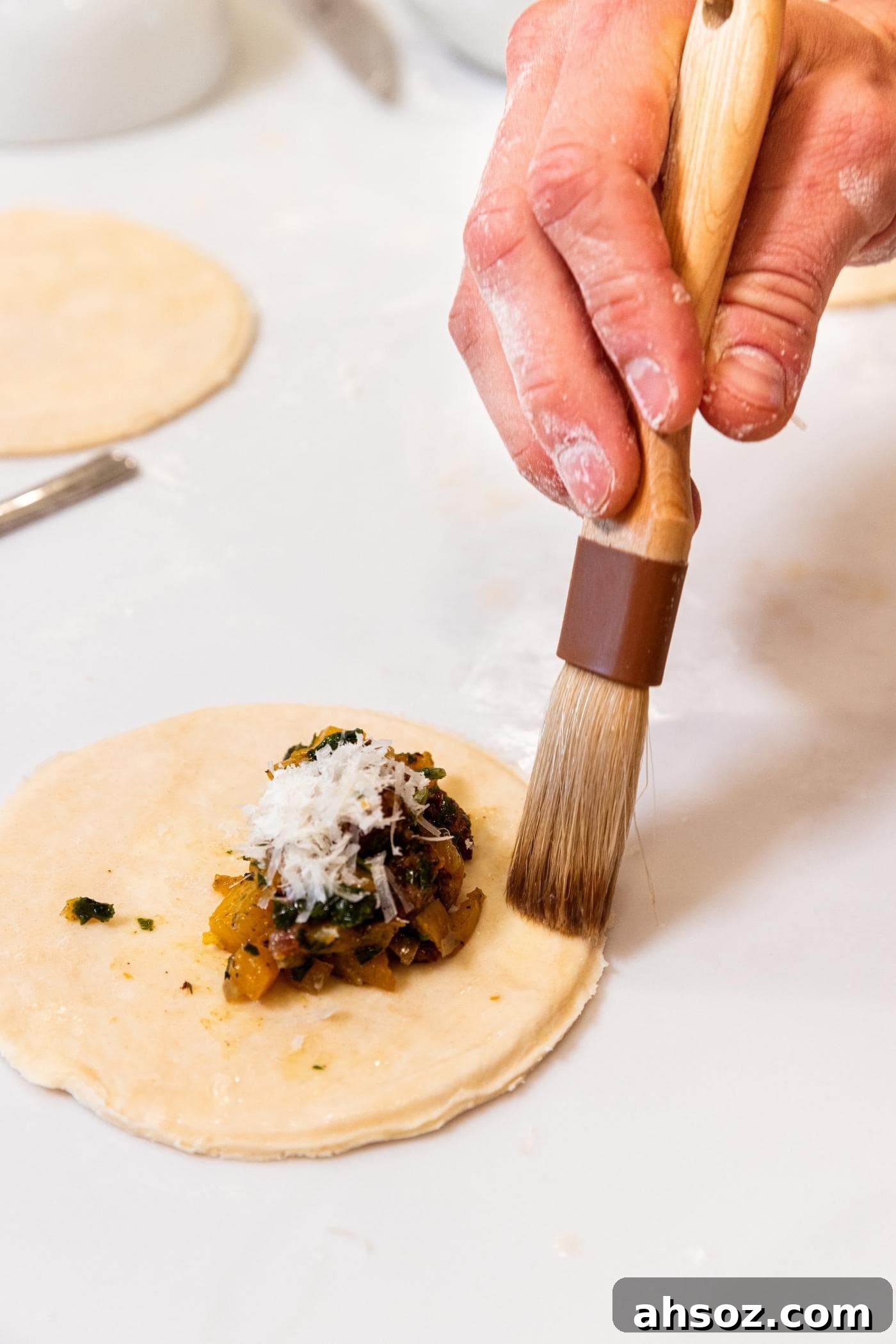 Hand using brush to use an egg wash to brush on one side of a circle of dough sitting on white countertop with small dollop of butternut squash and sausage mixture with Parmesan cheese on top.