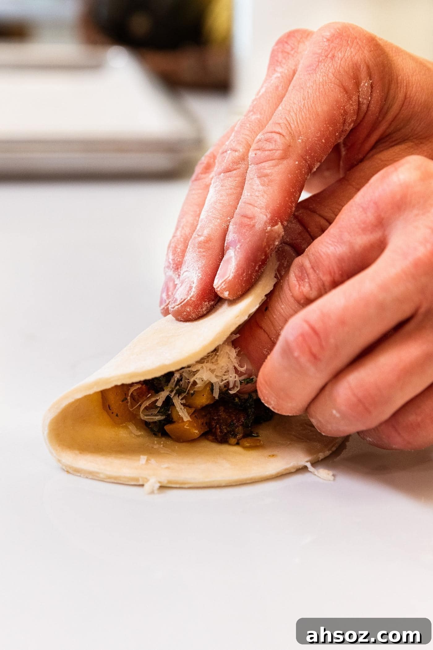 Two hands folding over half of a dough circle on top of a dollop of butternut squash and sausage mixture all sitting on white countertop.