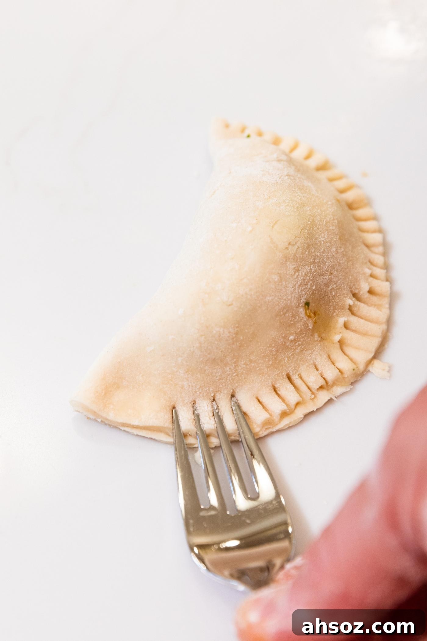Fork pressing tines down into dough of half moon shaped hand pie sitting on white countertop.