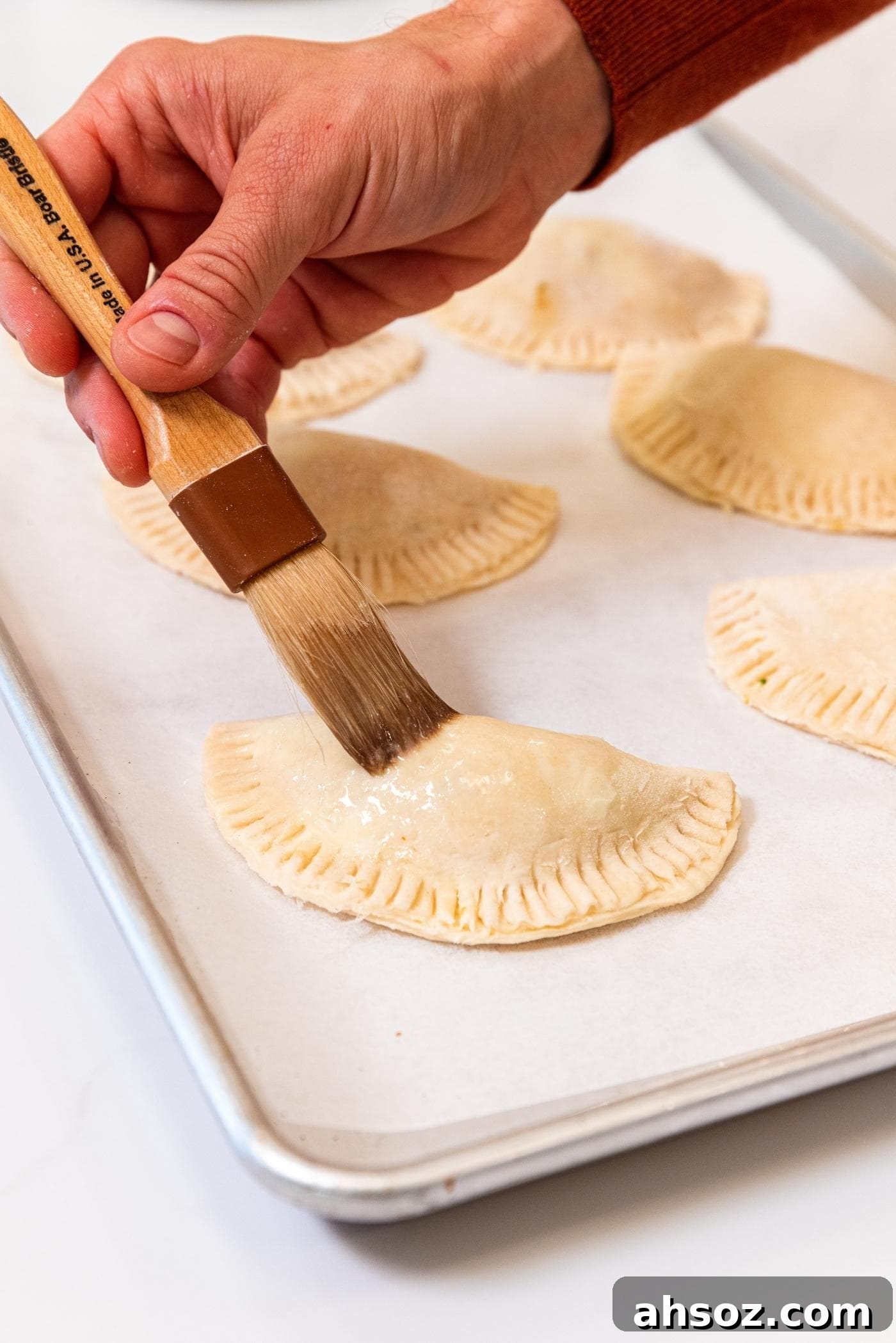 Hand using a brush to put an egg wash on the top of a half moon shaped hand pie sitting on a parchment-lined baking sheet with more hand pies in background.