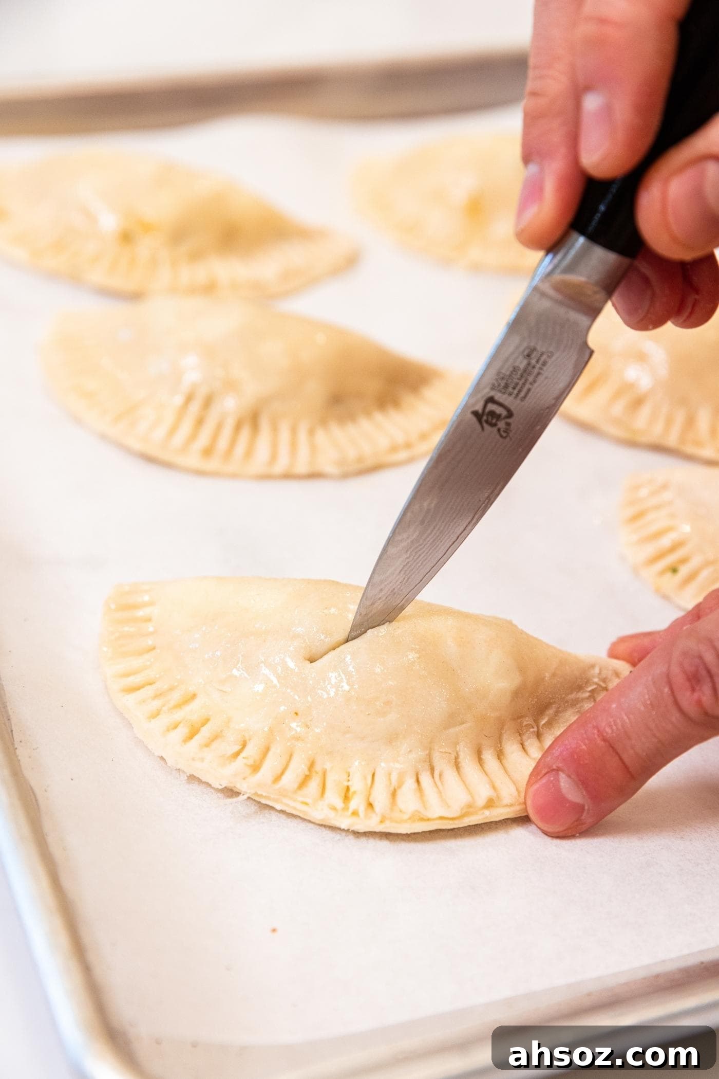 Hand using a knife to slice a vent hole in the top of a half shaped hand pie sitting on a parchment-lined baking sheet with more hand pies in the background.