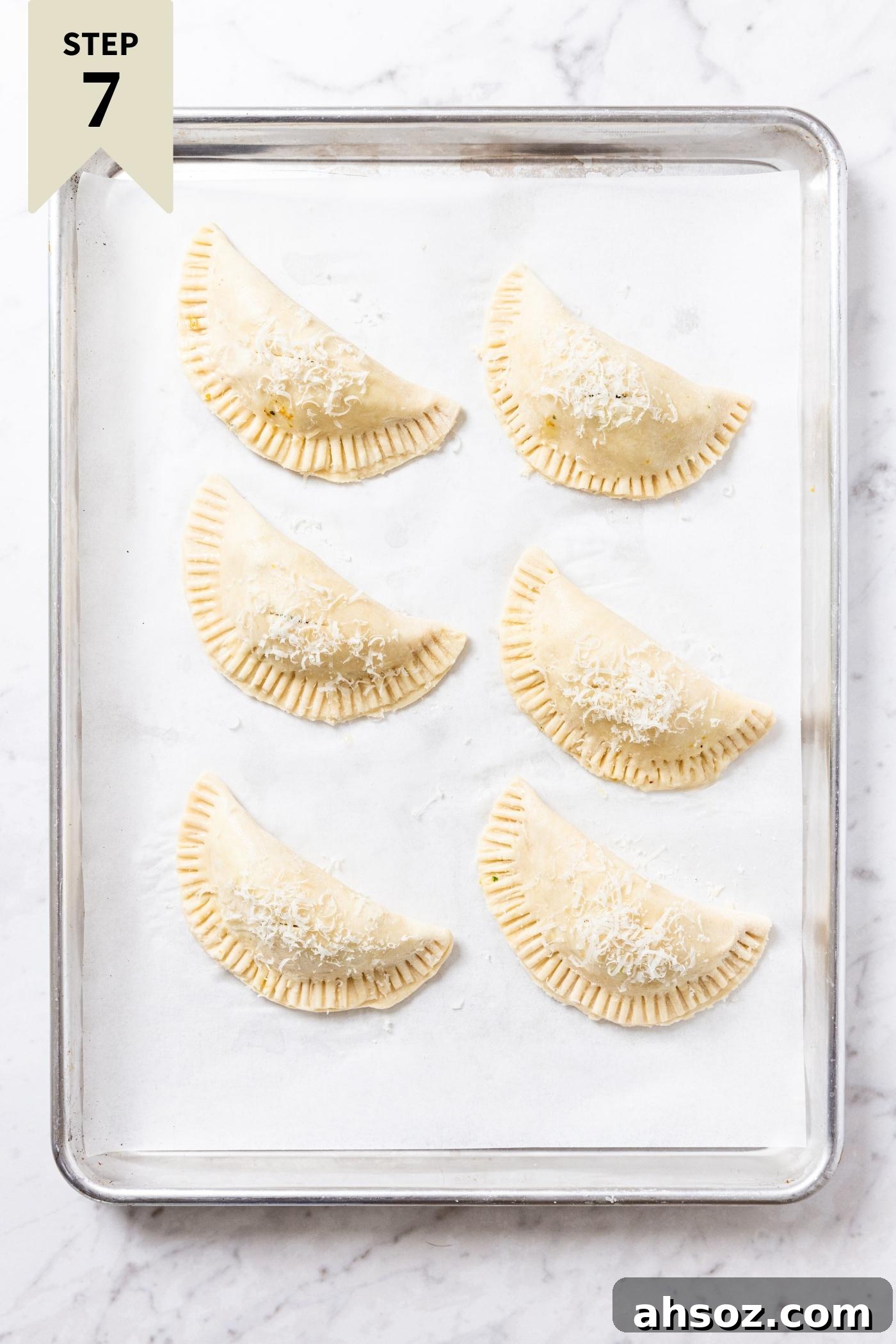 Unbaked half moon shaped hand pies sitting on a white parchment-lined baking sheet sitting on marble countertop before going into the oven to bake.