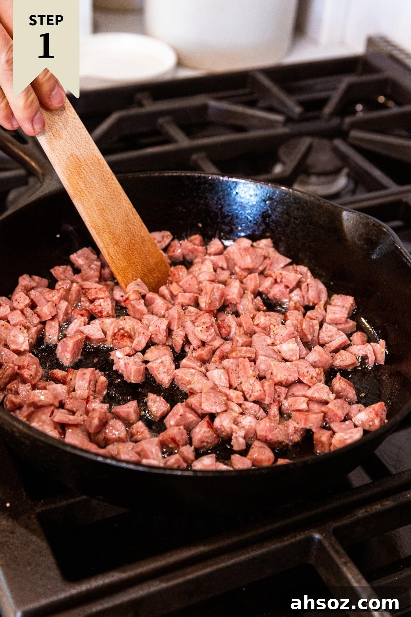 Wooden spatula stirring sausage cut into small diced squares in black cast iron skillet on stovetop.