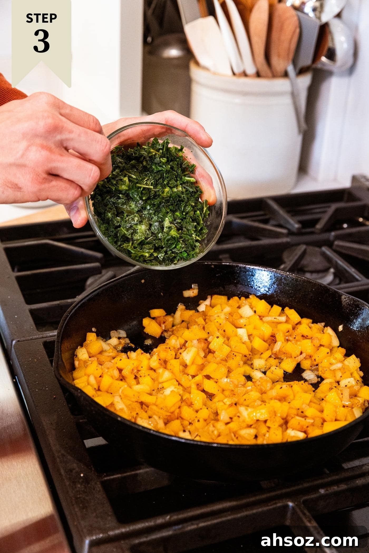 Hand holding small glass bowl filled with frozen kale about to pour it into a cast iron skillet on stovetop filled with cooked butternut squash pieces.