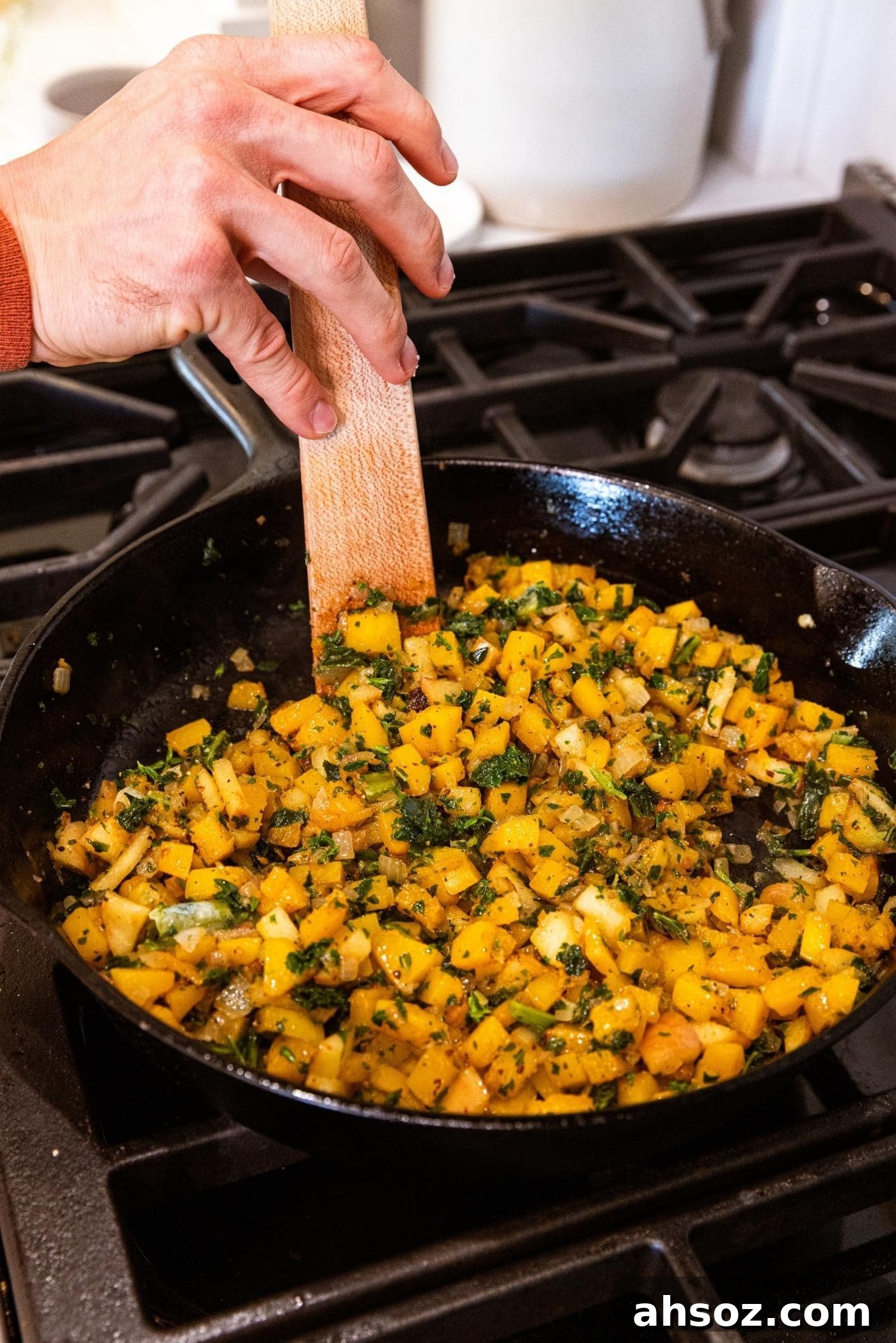 Hand using wooden spatula to mix together pieces of butternut squash and kale in black cast iron skillet on stovetop.