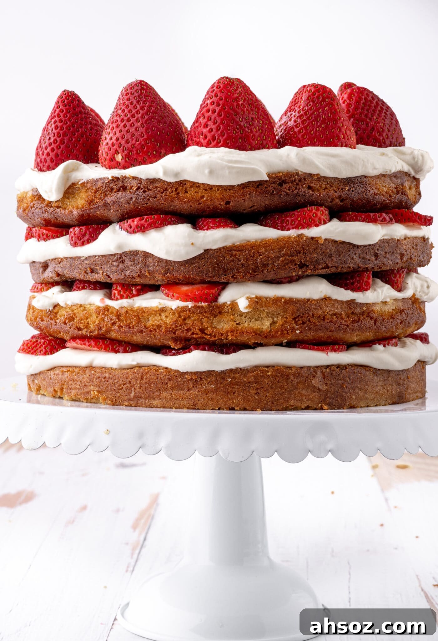 A four layer low carb strawberry shortcake sliced with fresh strawberries and whipped cream on a white ruffled cake stand.