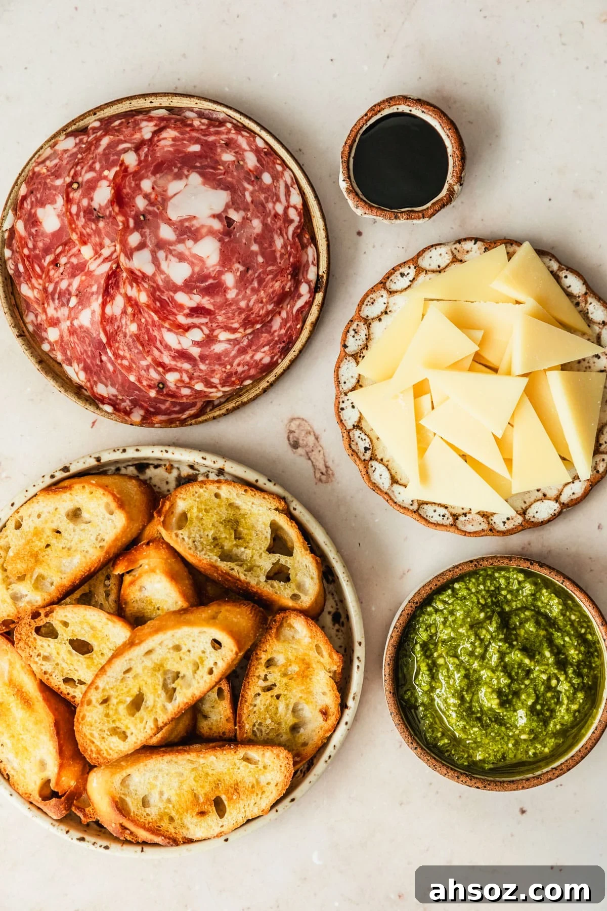 Brown and white bowls of soppressata, parmesan, pesto, crostini, and balsamic glaze on a tan counter.