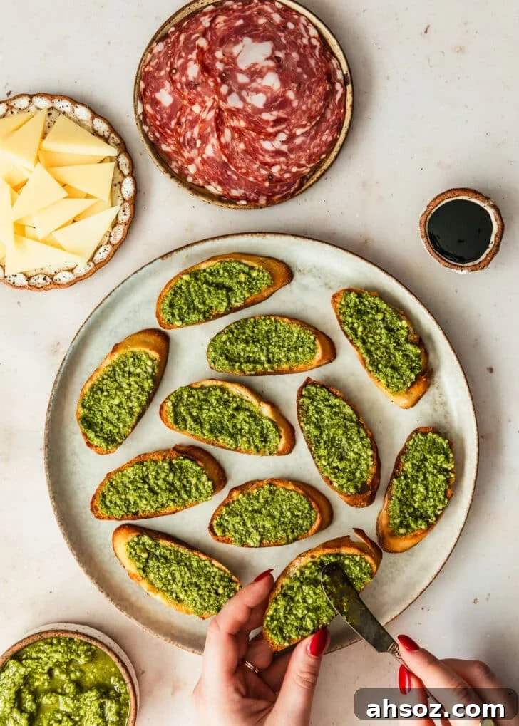A hand spreading basil sauce on toasts on a grey plate next to brown bowls of soppressata, parmesan, and balsamic glaze with a tan background.