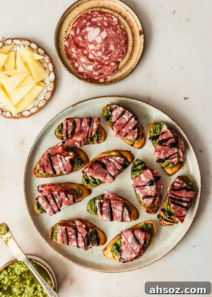 A grey platter of toasts with salami on a tan counter next to brown bowls of green sauce, parmesan, and salami.