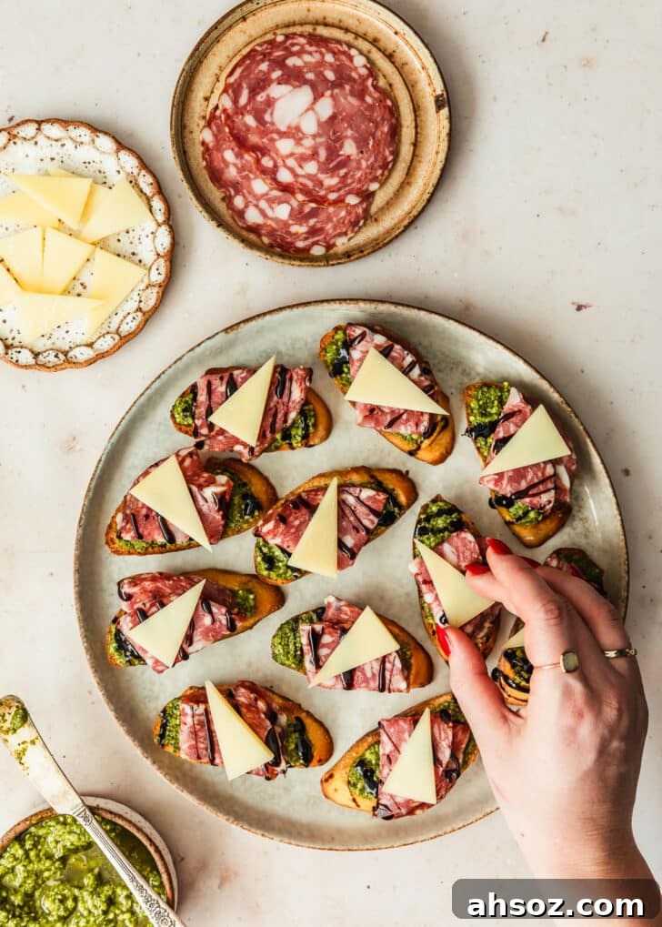 A hand placing slices of parmesan on a plate of soppressata pesto crostini next to brown bowls of pesto, soppressata, and parmesan with a tan marble background.