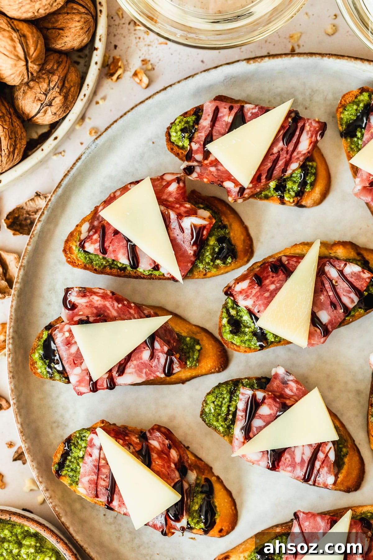 Soppressata, parmesan, and pesto crostini on a grey plate next to glasses of white wine, a white bowl of walnuts, and a brown bowl of pesto with a tan background.