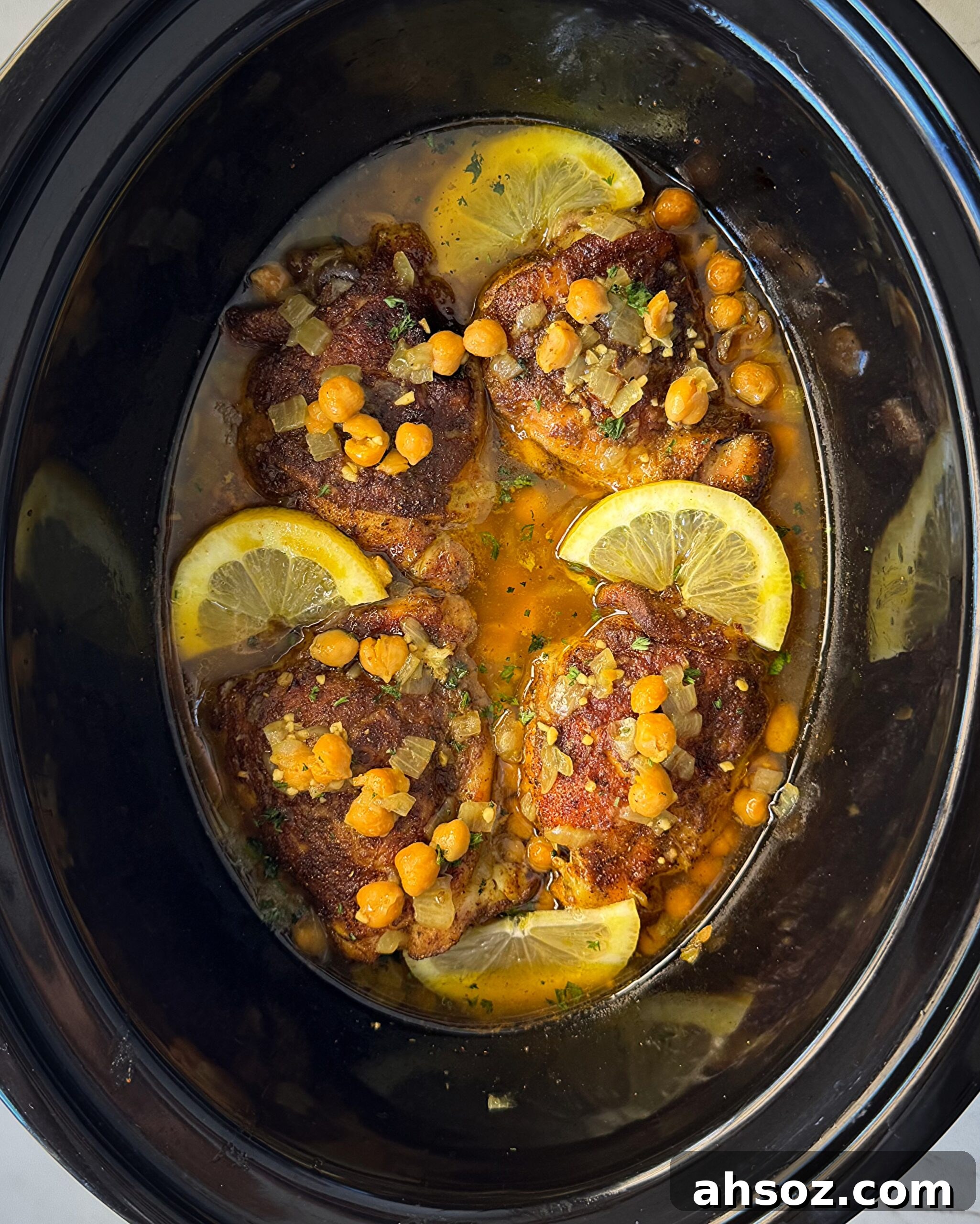 overhead shot of cooked North African chicken in a black slow cooker garnished with fresh parsley & sliced lemon