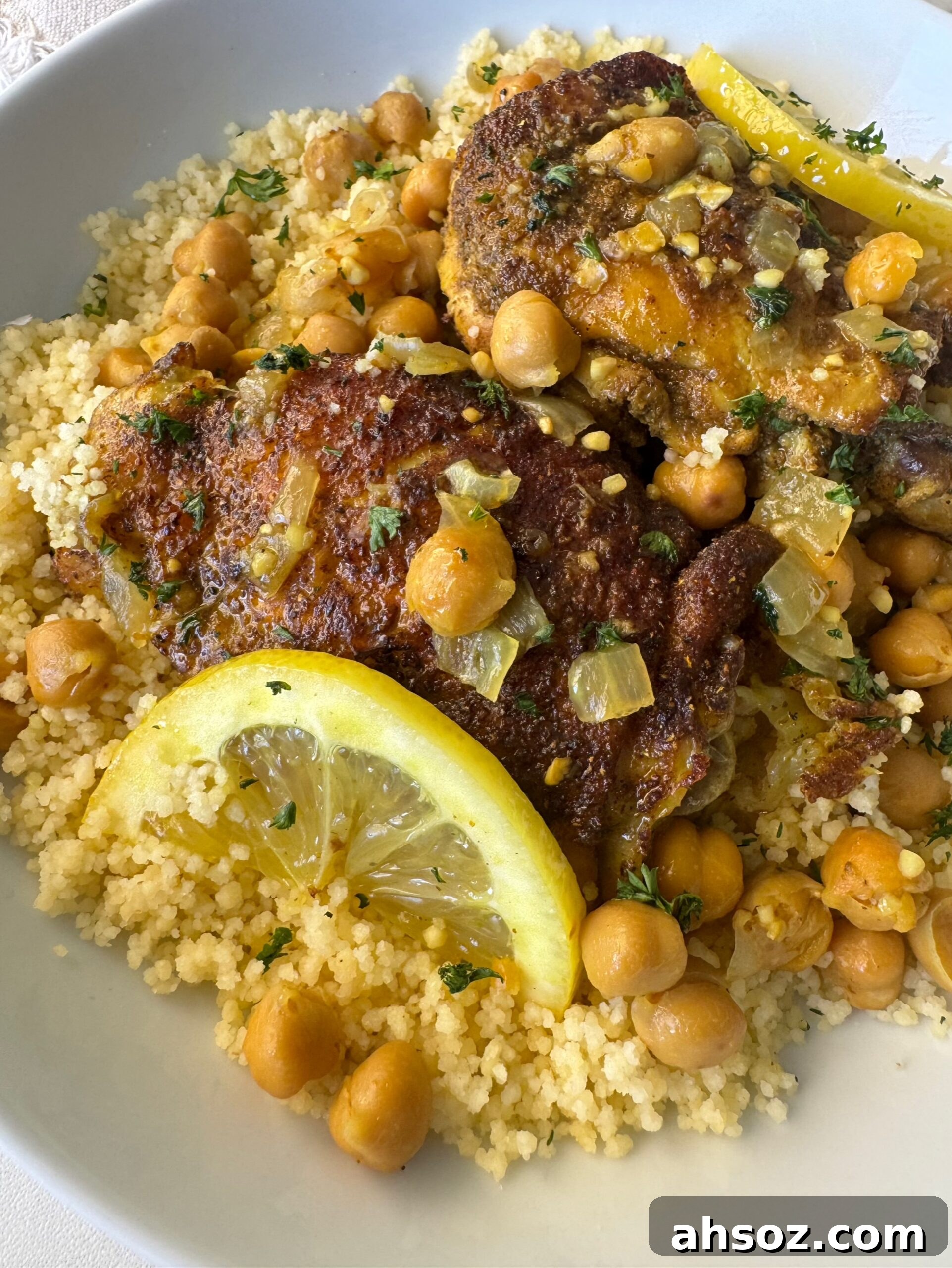 close up shot of cooked North African chicken on a white plate garnished with fresh parsley & sliced lemon served over couscous
